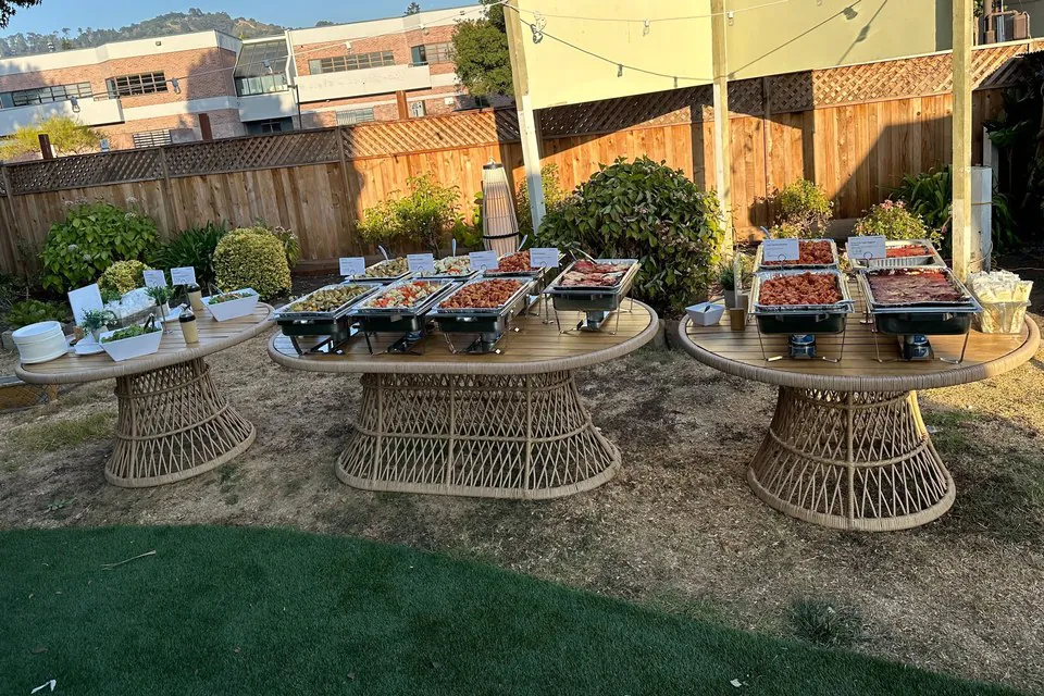 Outdoor offsite buffet on three round woven tables with chafing dishes of vegetables and grilled meats in a backyard venue at golden hour