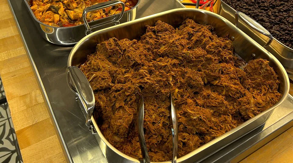 Deep steel tray of shredded barbacoa with serving tongs, beside black beans and a saucy mushroom tray on an office counter
