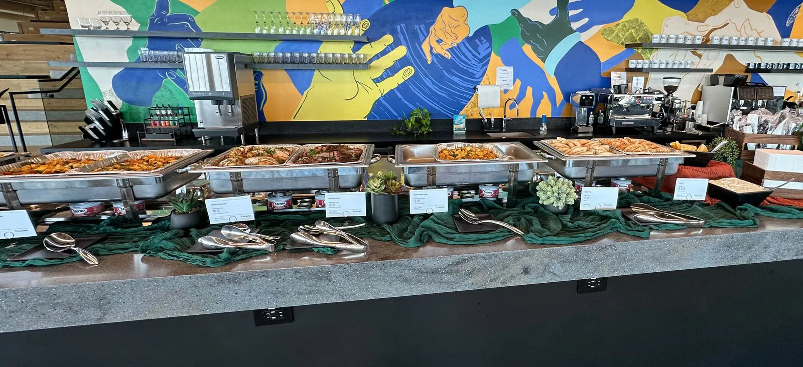 Office buffet line of full chafing trays with roasted vegetables, sliced meats and pastries along a stone counter under a colorful mural
