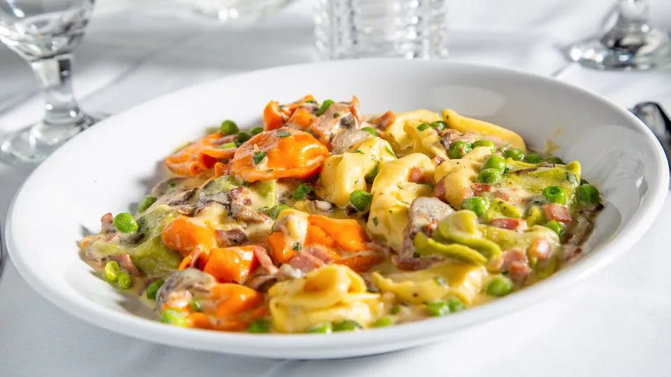 Tri-color tortellini in cream sauce with peas, prosciutto and mushrooms in a white bowl on a linen tablecloth