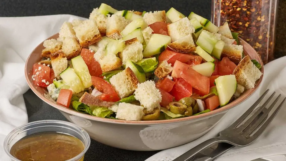 Chopped Italian salad with tomatoes, cucumbers, olives, gorgonzola and torn bread in a bowl with dressing in a cup on the side