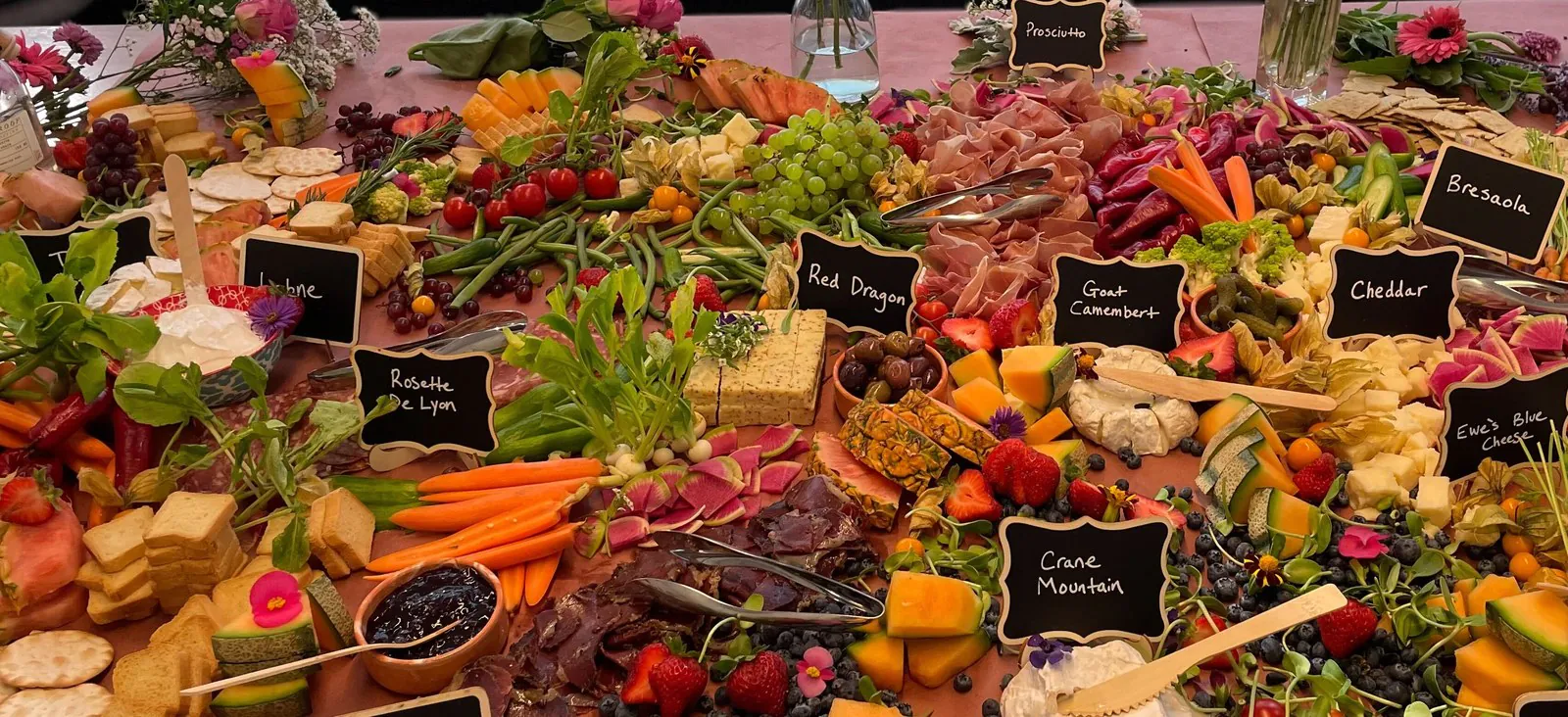 Long grazing table covered with cheeses, cured meats, crackers, fruit and vegetables, each item marked with a small handwritten chalkboard label