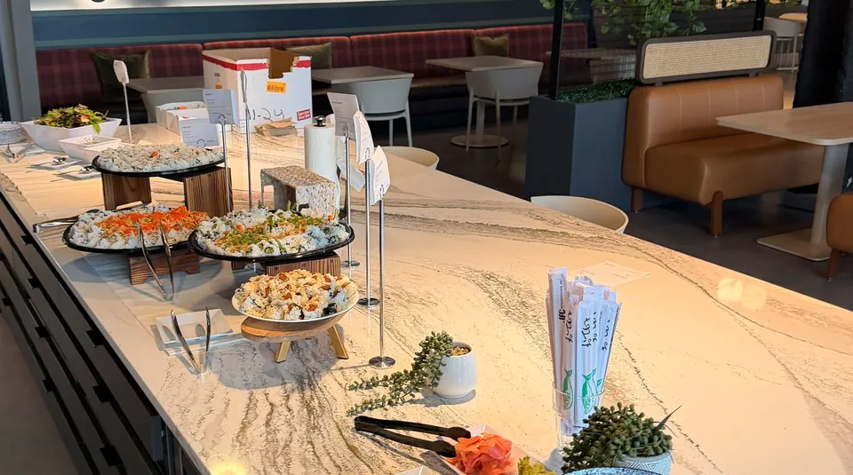 Long stone counter in an office lounge holding platters of sushi and salads with small printed cards, with banquette seating and cafe tables behind