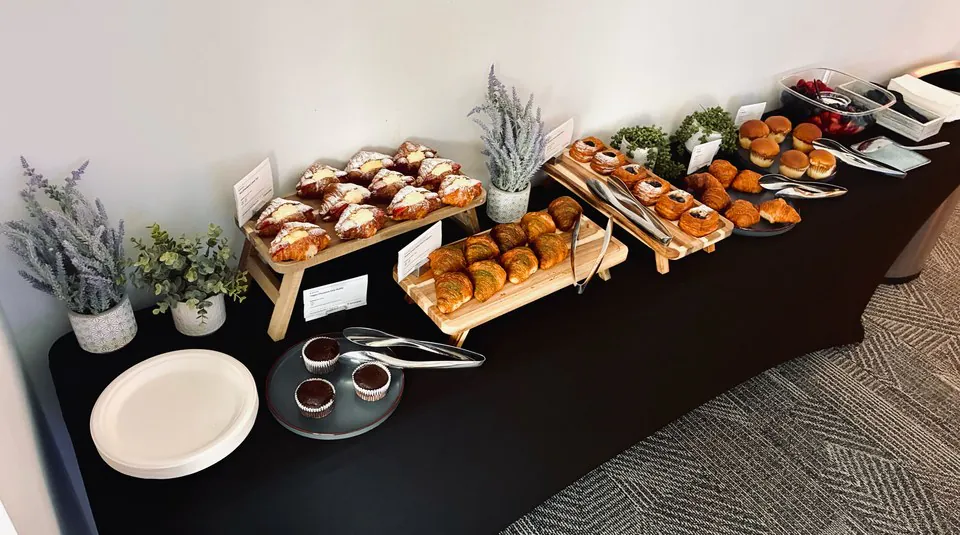 Pastry spread of croissants, danishes and muffins on wooden risers along a black-draped table with printed cards and small plants