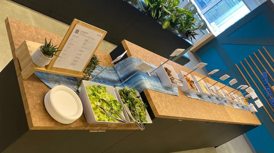 Self-serve salad station on a wooden counter with white bins of greens and toppings, each marked with a small card, beside plates and a menu stand