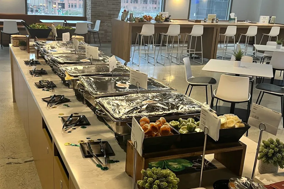 Long office buffet line with chafing trays, serving tongs, plates and a condiment bar along a marble counter