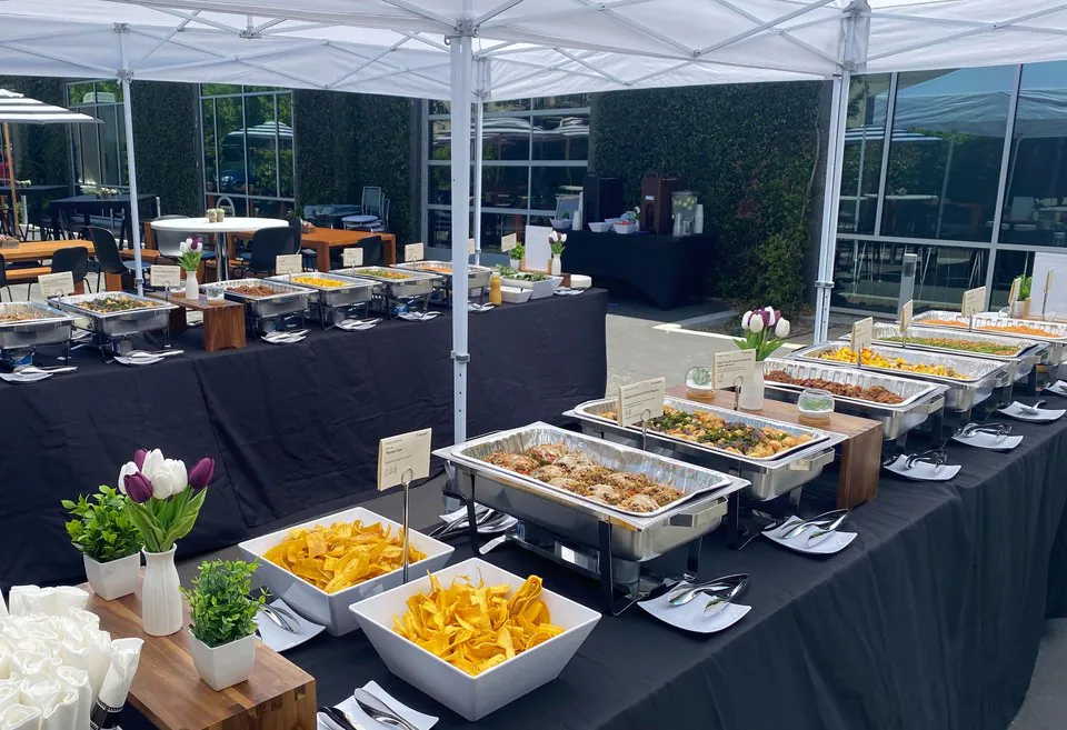 Long outdoor catering buffet under white tents with chafing dishes and menu cards