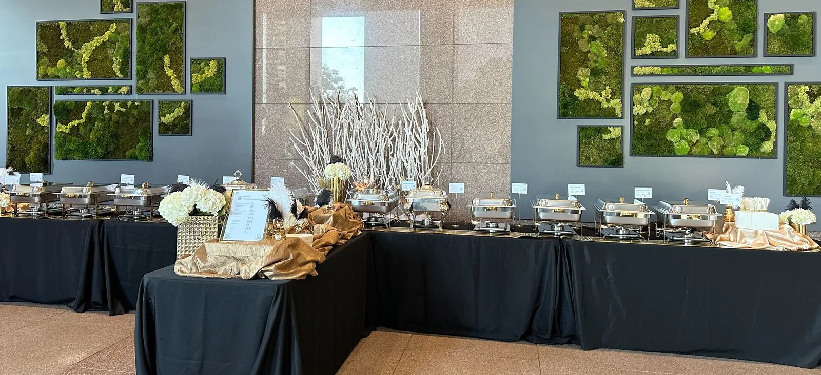 Office catering buffet with rows of chafing dishes on black draped tables under moss wall art