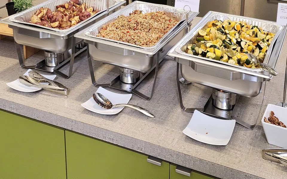 Three chafing trays of roasted potatoes, grain pilaf, and fire-grilled squash on an office kitchen counter
