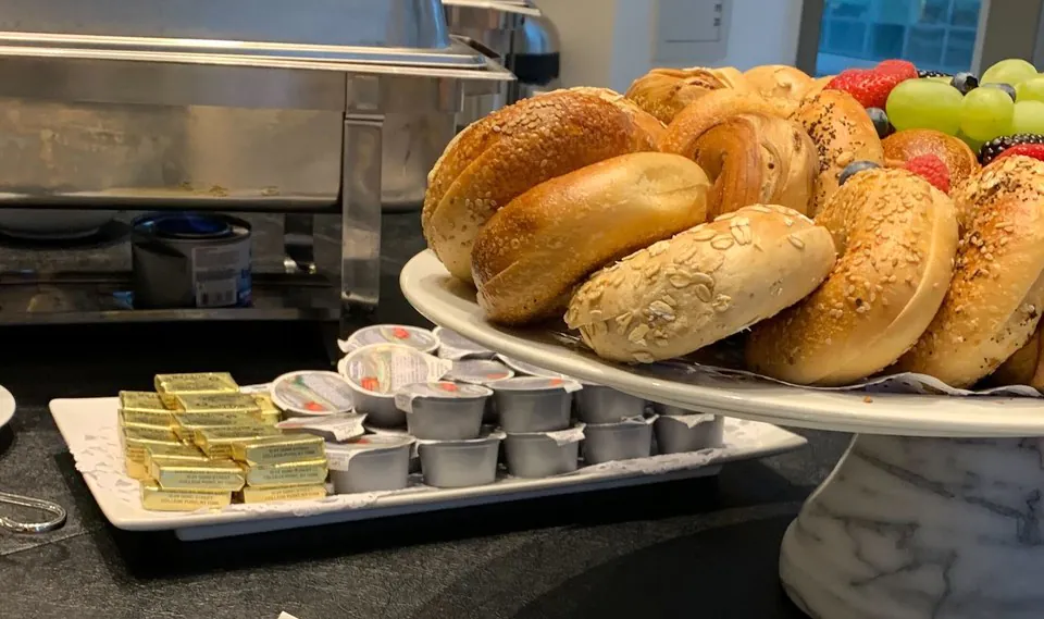 Office breakfast tray of assorted bagels and fruit beside butter and cream cheese cups on a dark counter