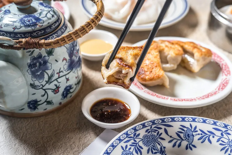 Chopsticks lifting a pan-fried dumpling above a dish of chili sauce, with a plate of pan-fried dumplings, steamed rice rolls and a blue and white teapot on a dim sum table