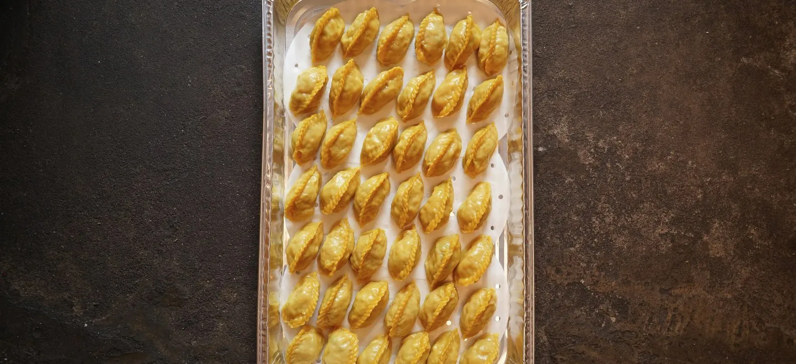 Aluminium catering tray filled with rows of pleated golden dumplings on a dark stone surface