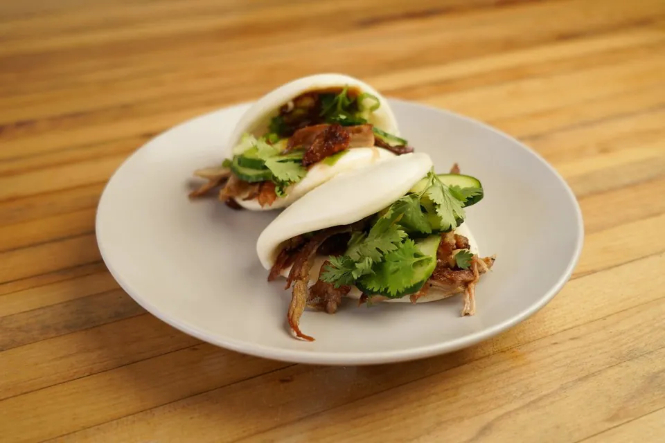 Two Peking duck buns filled with shredded duck, cucumber and cilantro on a white plate on a wooden table