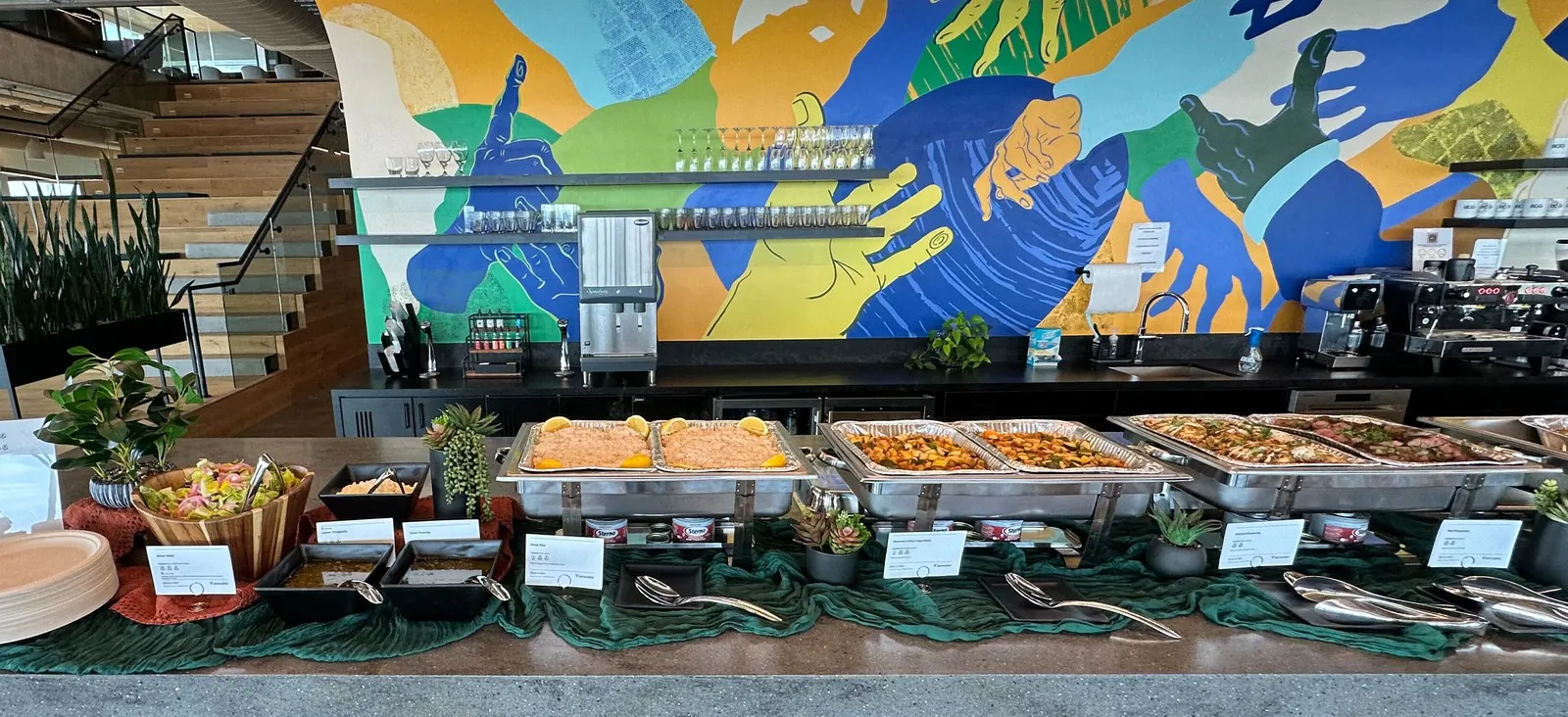 Office buffet line of foil chafing trays with rice,roasted vegetables and grilled mains along a stone counter under a colorfulhand mural