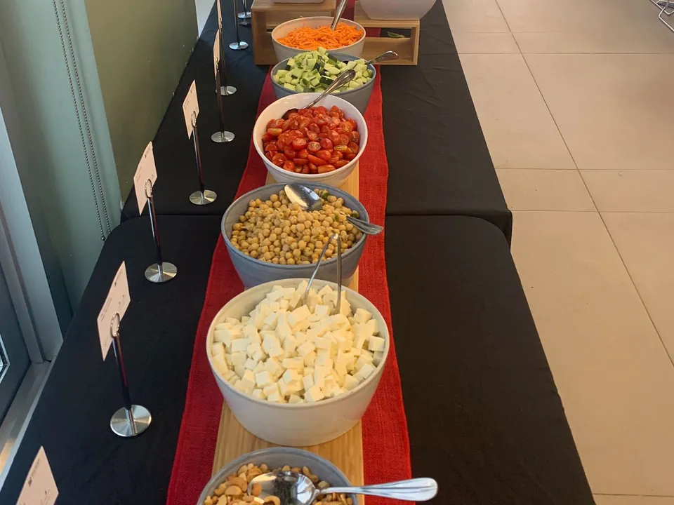 Linear build station with bowls of cashews, dicedmozzarella, chickpeas, cherry tomatoes, cucumber and shredded carrot on a redand black runner