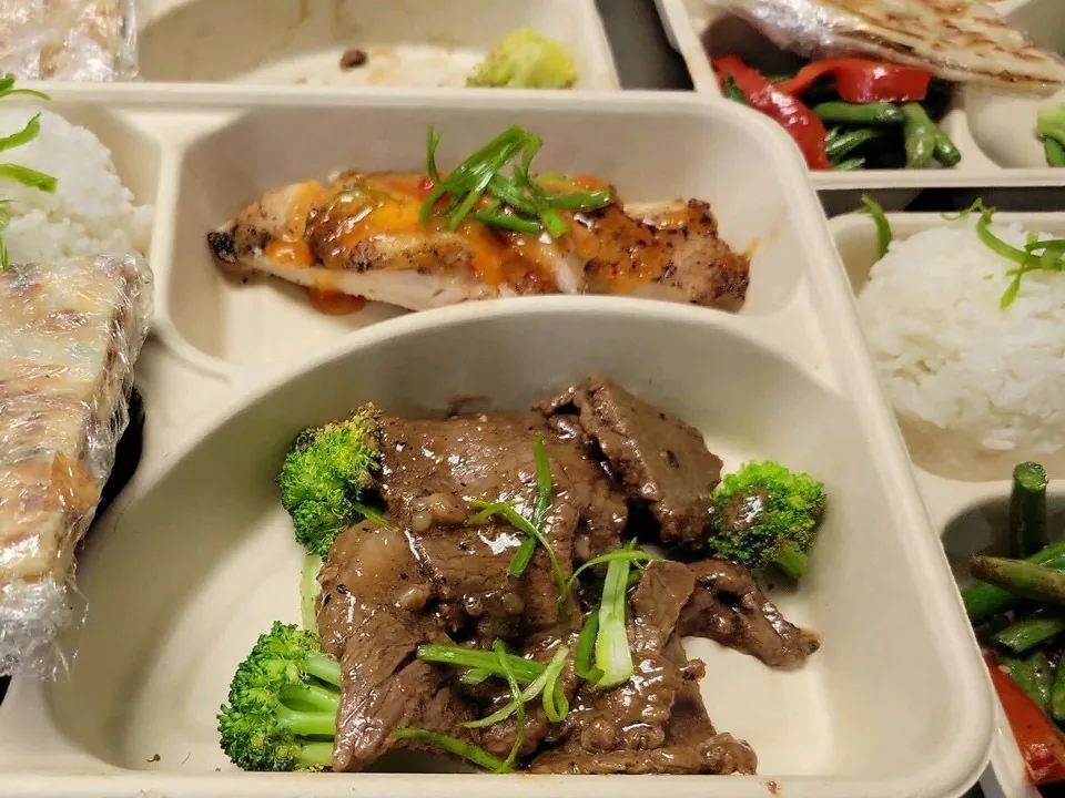 Compartmented individual meal trays with steamedrice, beef and broccoli, grilled chicken and green beans lined up on a counter