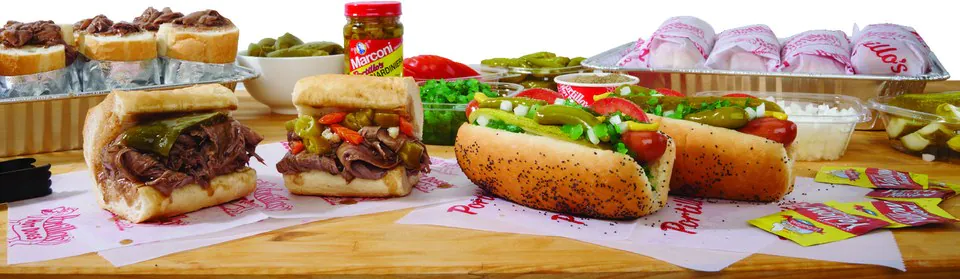 Portillo's Italian beef sandwiches and Chicago-style hot dogs on a wooden board beside tubs of chopped onions, relish, tomatoes and giardiniera, with wrapped sandwiches in a foil tray behind