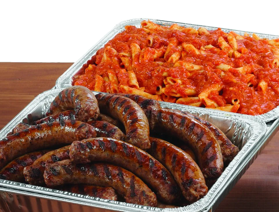 Two foil catering pans on a wooden table, one filled with mostaccioli in red sauce and one with char-grilled Italian sausages