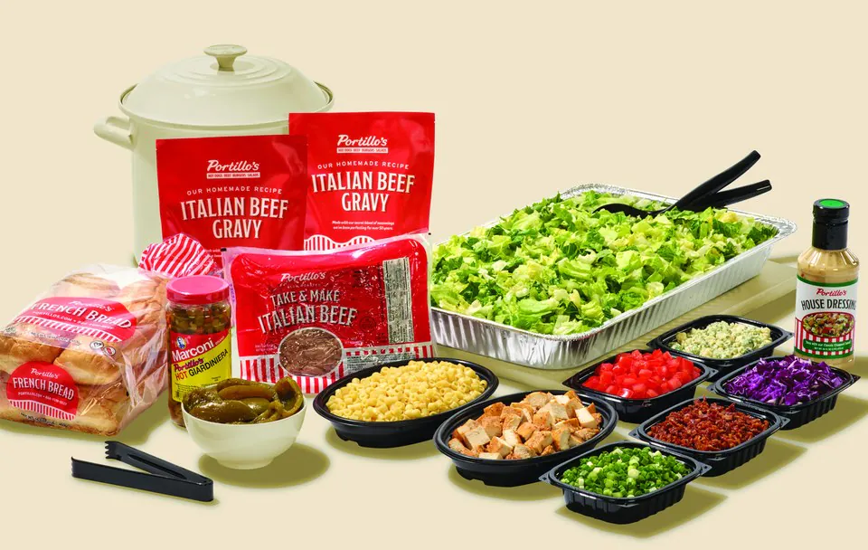 A Portillo's Take and Make kit laid out: sealed Italian beef, two pouches of gravy, a bag of French bread rolls, a jar of hot giardiniera, a foil pan of chopped romaine and eight separate tubs of salad toppings