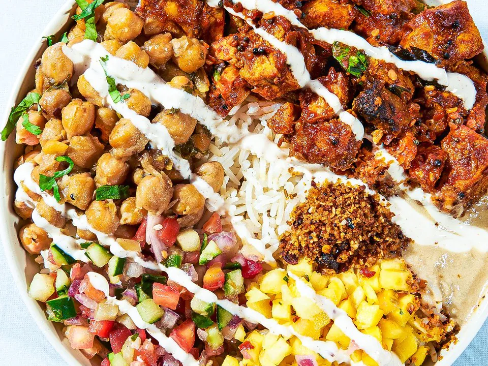 Overhead view of a Rasa bowl with chickpeas,chicken tikka, kachumber salad, mango salsa and basmati rice kept in separatewedges under a yogurt drizzle