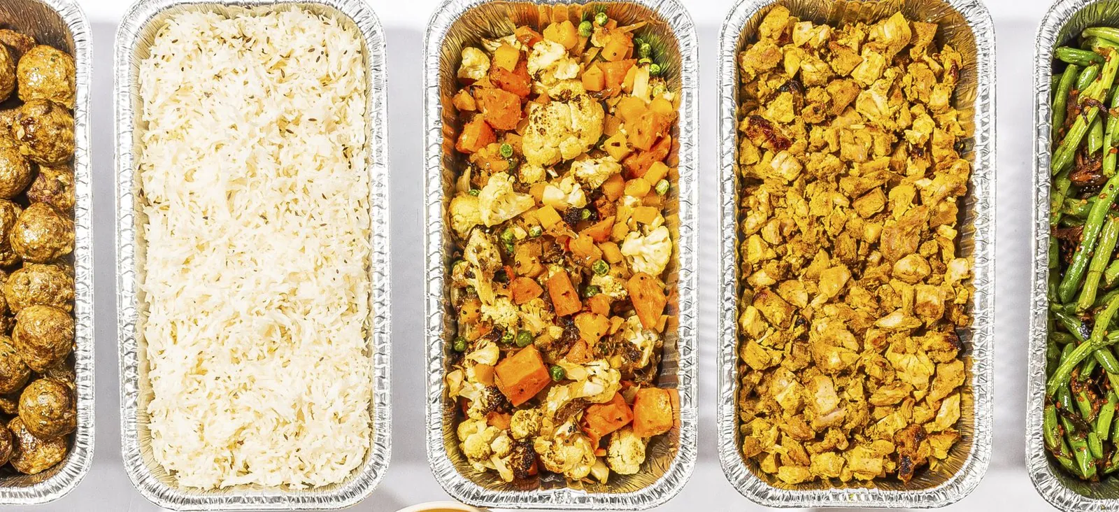 Five foil catering trays of lamb kebab, basmatirice, roasted cauliflower and potato, chicken tikka and green beans in a row ona white surface