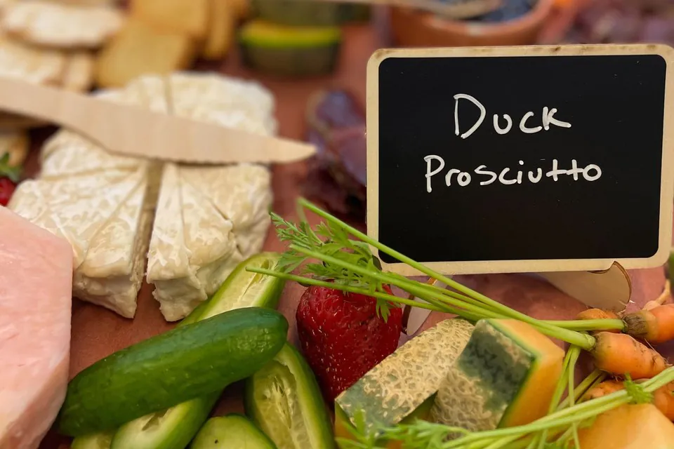 Handwritten chalkboard label reading Duck Prosciutto beside cheese, cucumber and cut fruit on a board