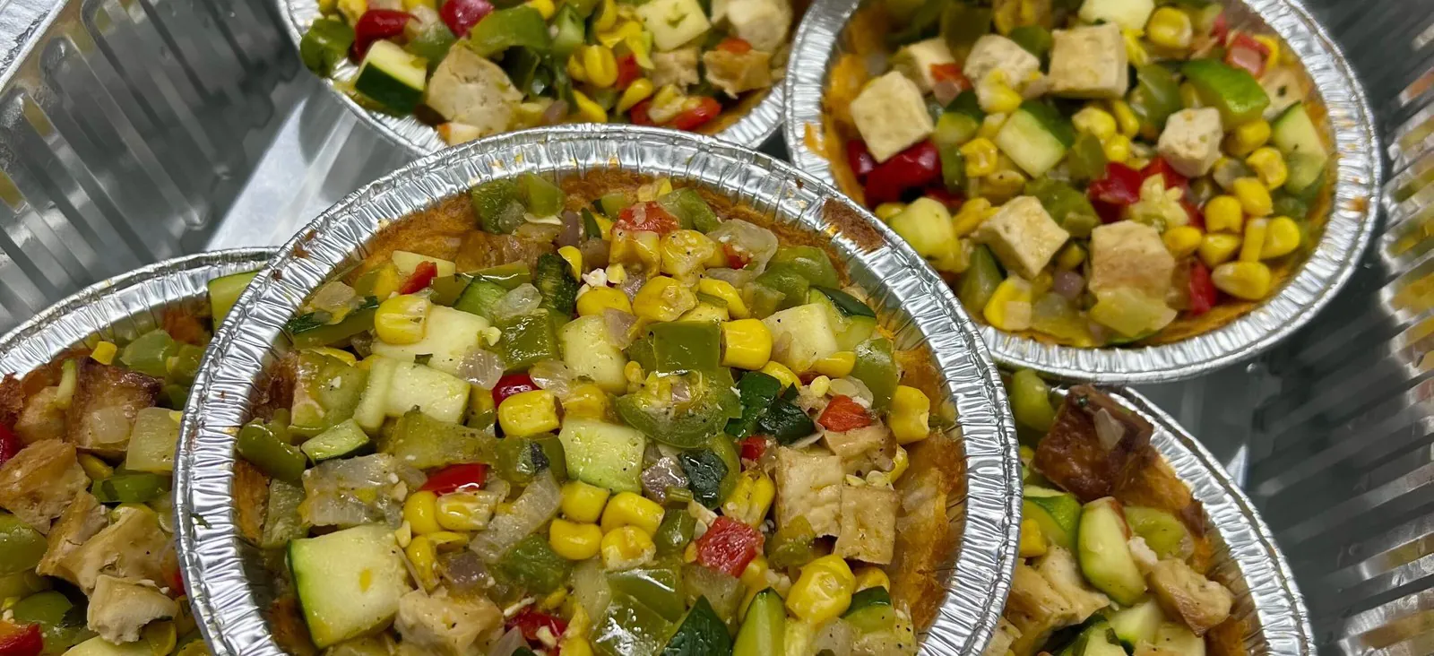 Individual foil bowls of corn, zucchini, peppers, and chicken portioned one per person in a catering tray