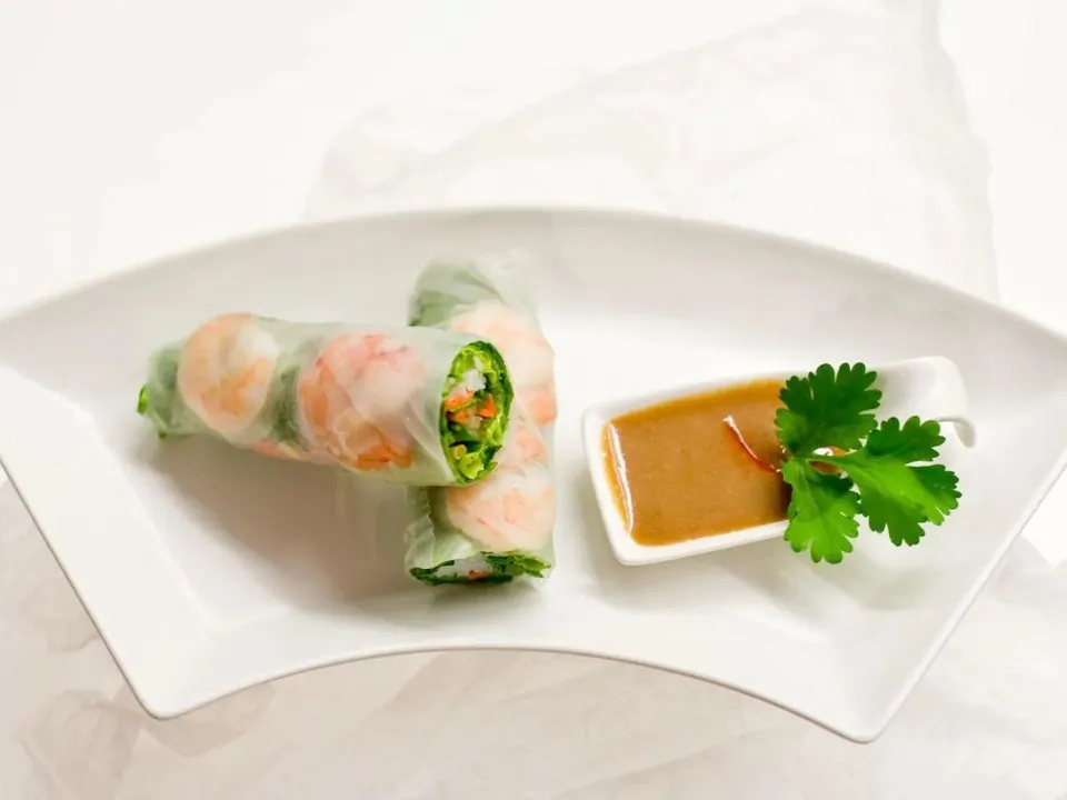 Fresh Vietnamese shrimp spring rolls on a whiteplate beside a small dish of peanut dipping sauce and a sprig of cilantro