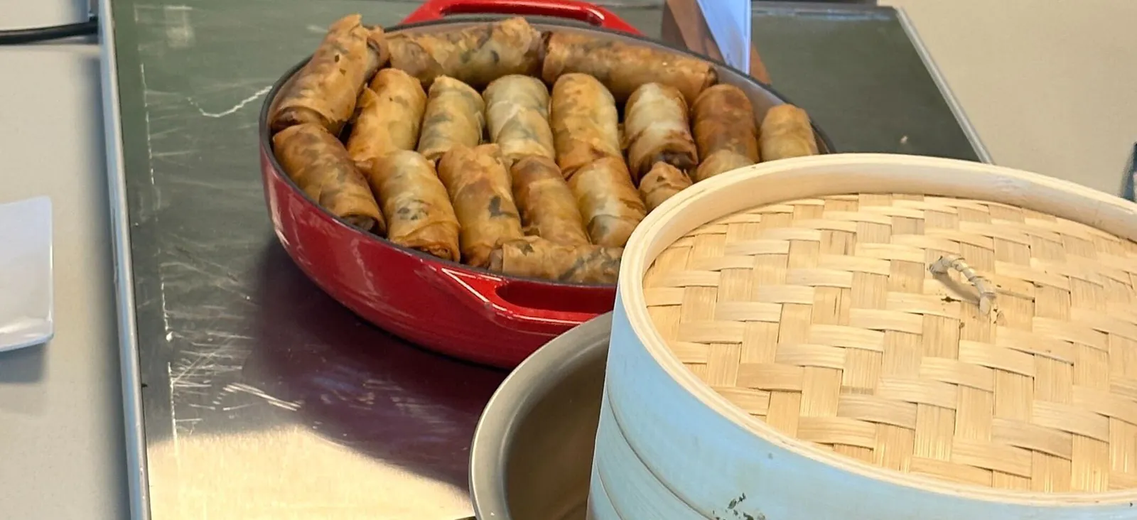 Fried Vietnamese spring rolls in a red enamel panon an office buffet warmer, with a bamboo steamer alongside