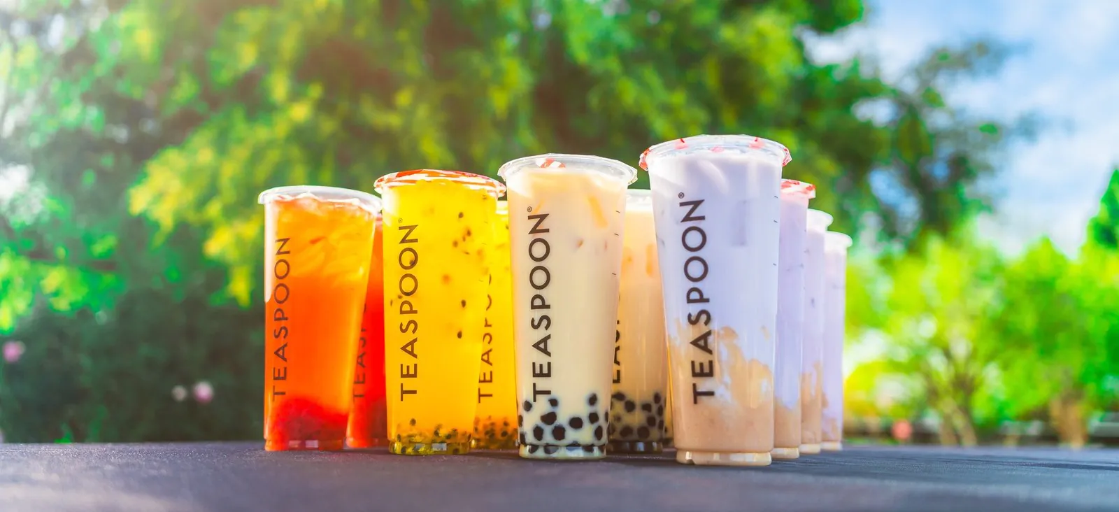 Row of sealed Teaspoon bubble tea cups in orange, mango, milk tea, taro and strawberry, lined up outdoors with tapioca pearls visible at the bottom
