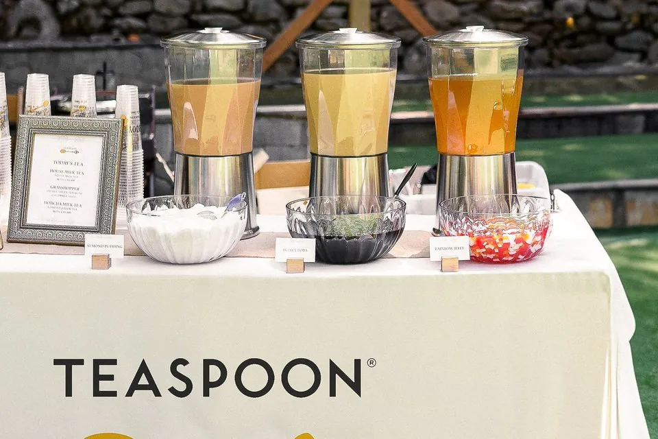 Teaspoon on-site catering table with three tea dispensers, labeled glass bowls of almond pudding, honey boba and rainbow jelly, stacked cups and a branded table skirt