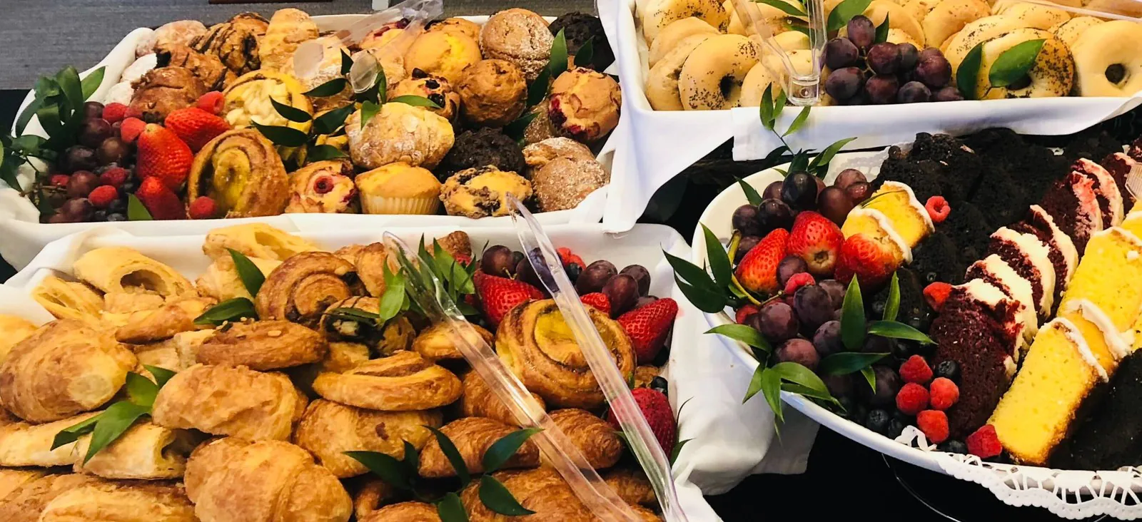 Office breakfast spread of croissants, danishes,muffins, bagels, sliced cakes and fresh berries laid out in white-lined trays