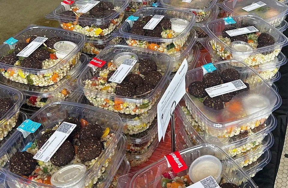 Rows of individually packaged grain salad meals inclear containers with labels, lined up along a catering table
