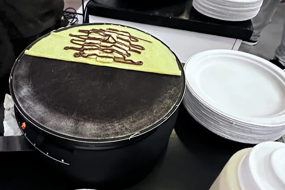 Two Crepes crepe cooking on the griddle Single crepe cooking on a round black griddle with chocolate sauce drizzled across it, stacked white plates beside the station