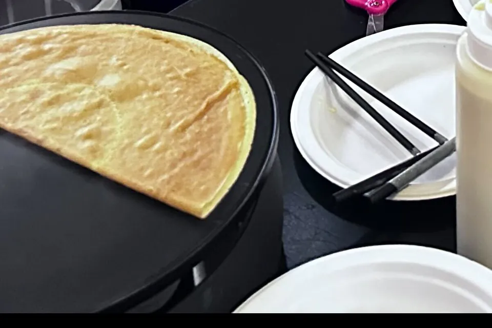 Two Crepes folded crepe at the cart Cooked crepe folded in half on a black griddle at the service window with spatulas, a paper plate and a batter bottle beside it