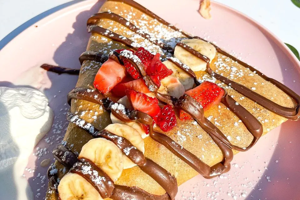 Two Crepes plated sweet crepe Sweet crepe on a plate topped with sliced strawberries, banana, chocolate hazelnut drizzle and powdered sugar