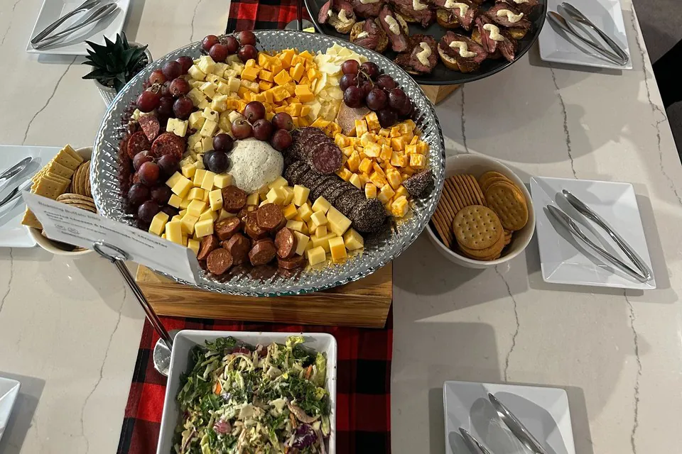 Round party tray of cubed cheddar, sliced sausage and grapes beside a bowl of crackers on a counter