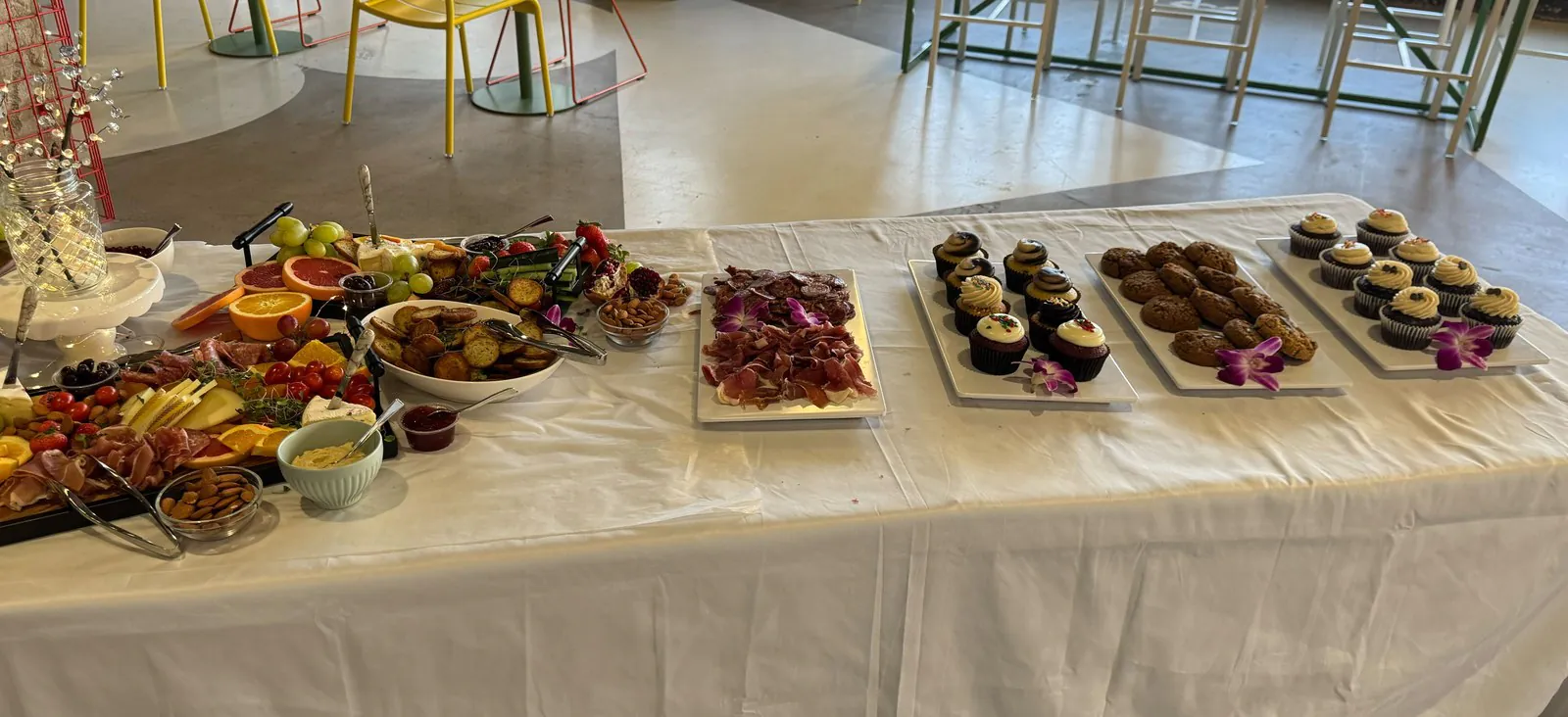 Long white catering table carrying a cured meat and cheese board, a sliced meat platter, and separate platters of cookies and cupcakes