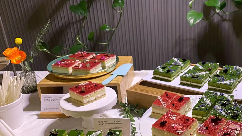 A footed white cake stand and wooden serving boards holding sliced layer cakes on a linen-covered display table with cake servers