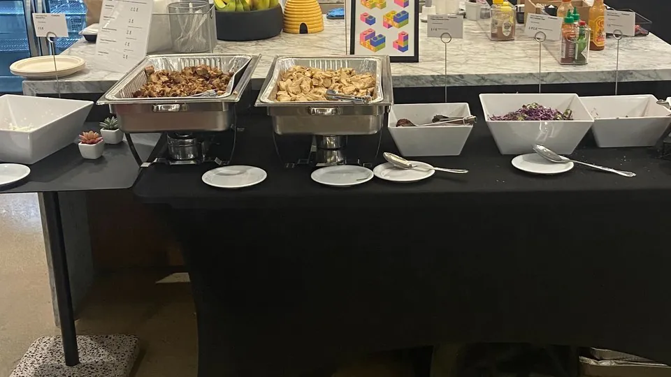 Stainless chafer frames holding foil pans beside white china bowls and side plates on a black-linen catering table in an office