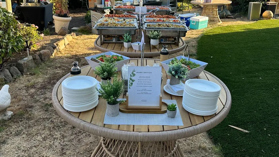 An outdoor catering station on a round timber table with two stacks of plates, a printed menu on a stand and chafers on wire stands behind