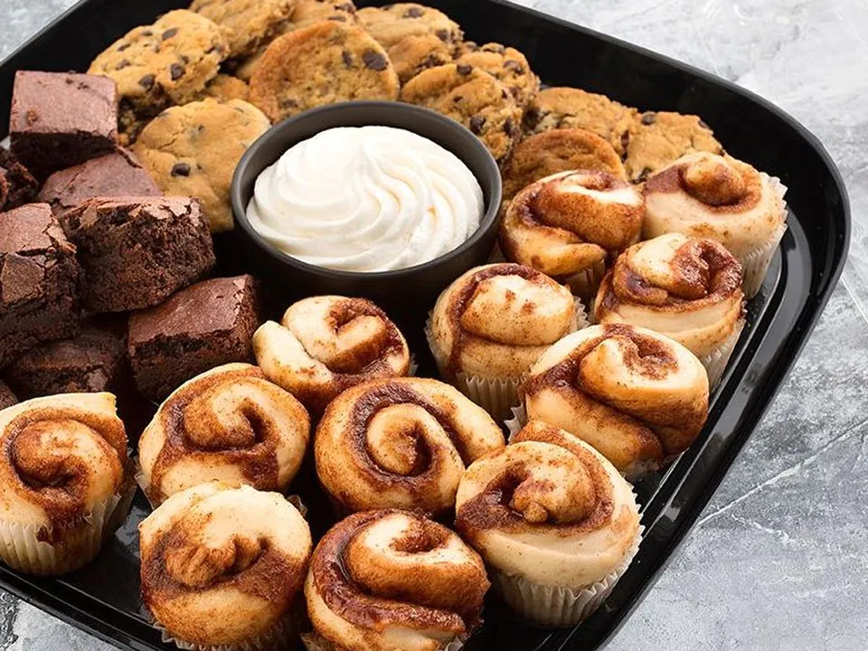 Black catering tray of mini cinnamon rolls, chocolate chip cookies, brownie squares and a cup of frosting