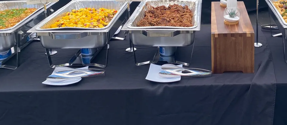 Foil half-pans of pulled meat and roasted vegetables in chafing frames on a draped catering table outdoors