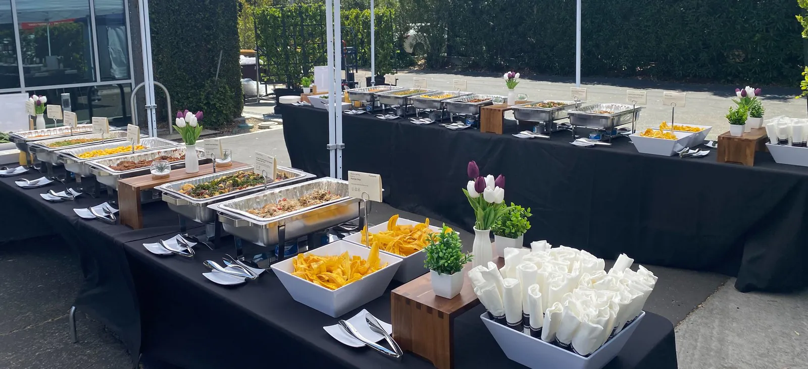 Parallel catering buffet lines with foil pans and chafing frames set up under white pop-up tents on asphalt