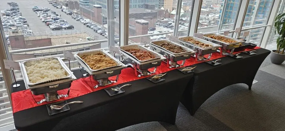 Row of foil chafing trays on a black draped catering table beside a window wall