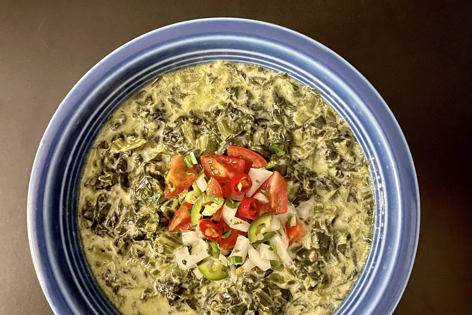 An overhead bowl of chopped greens in a pale coconut cream sauce topped with diced tomato, onion and green chili