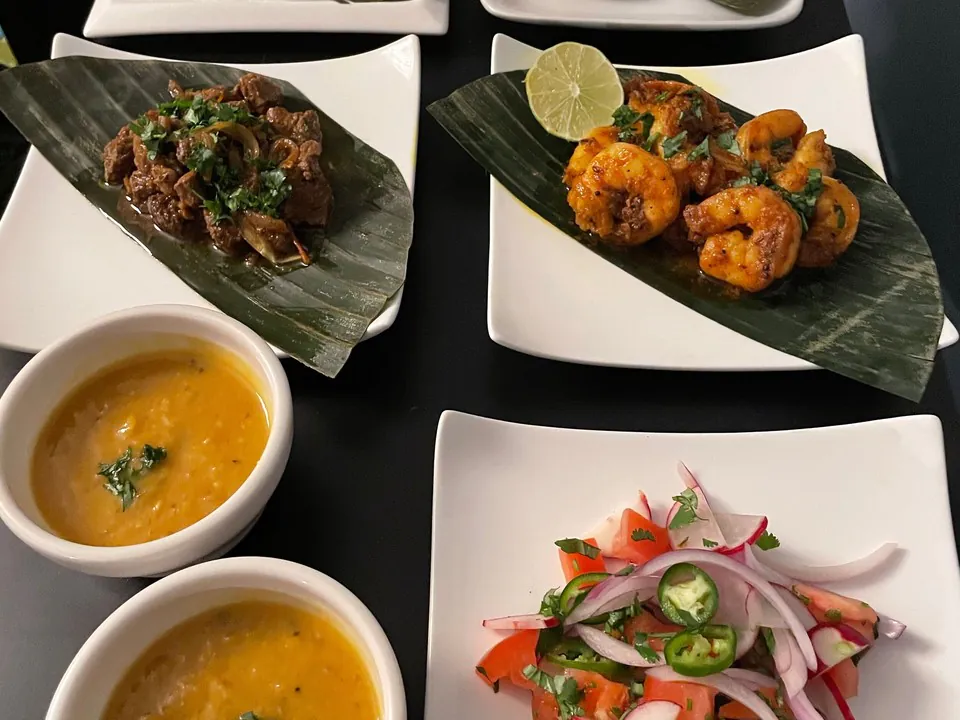 Rectangular white plates on a dark counter holding braised lamb and masala prawns on banana leaves, with two bowls of yellow dhal and a fresh salad