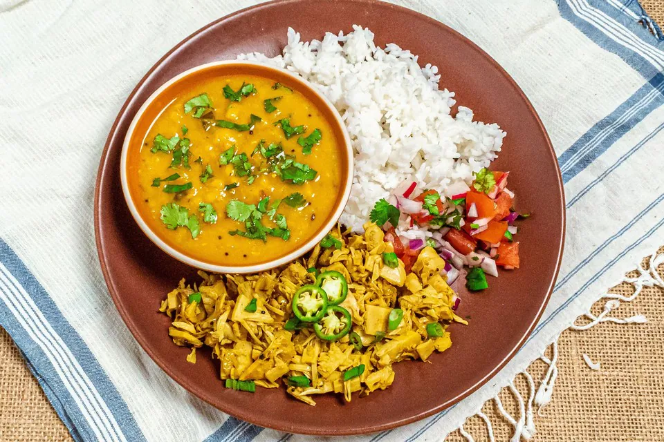 A terracotta plate with a bowl of yellow lentil dhal, white rice, shredded jackfruit and a fresh tomato and onion relish