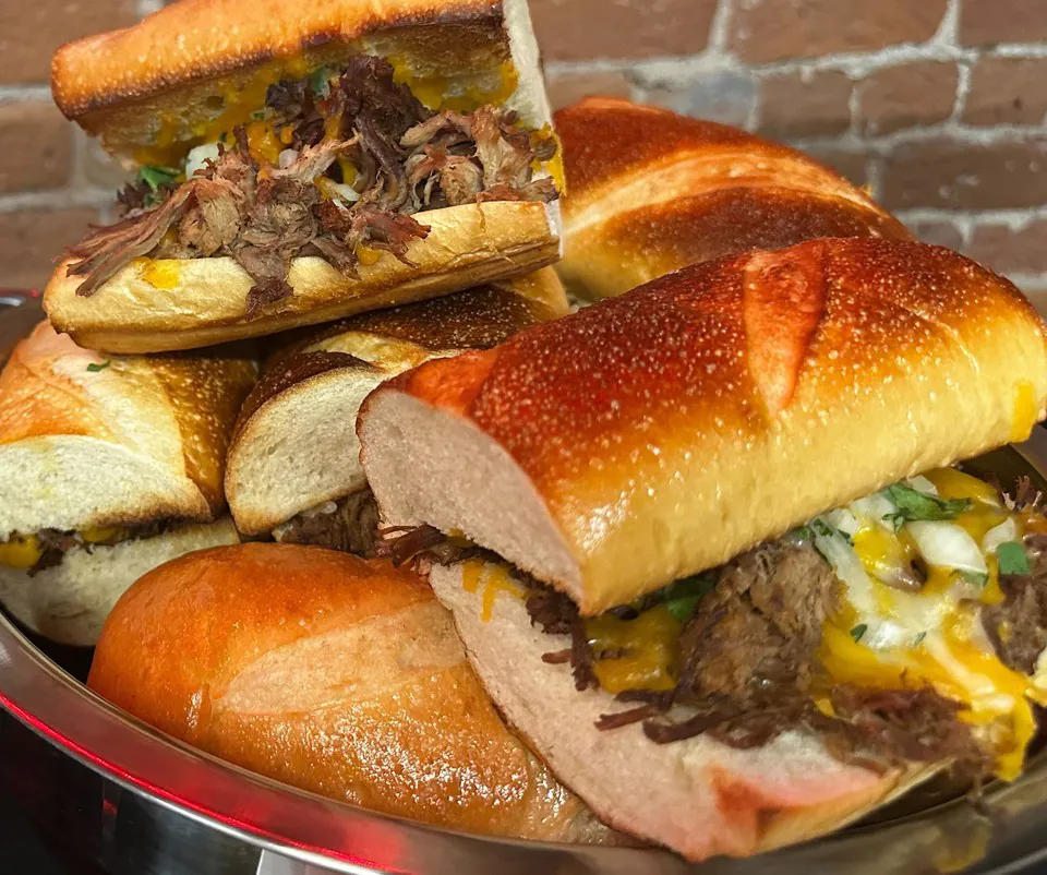 Stacked beef sandwiches on brioche rolls piled in a metal catering tray against an exposed brick wall