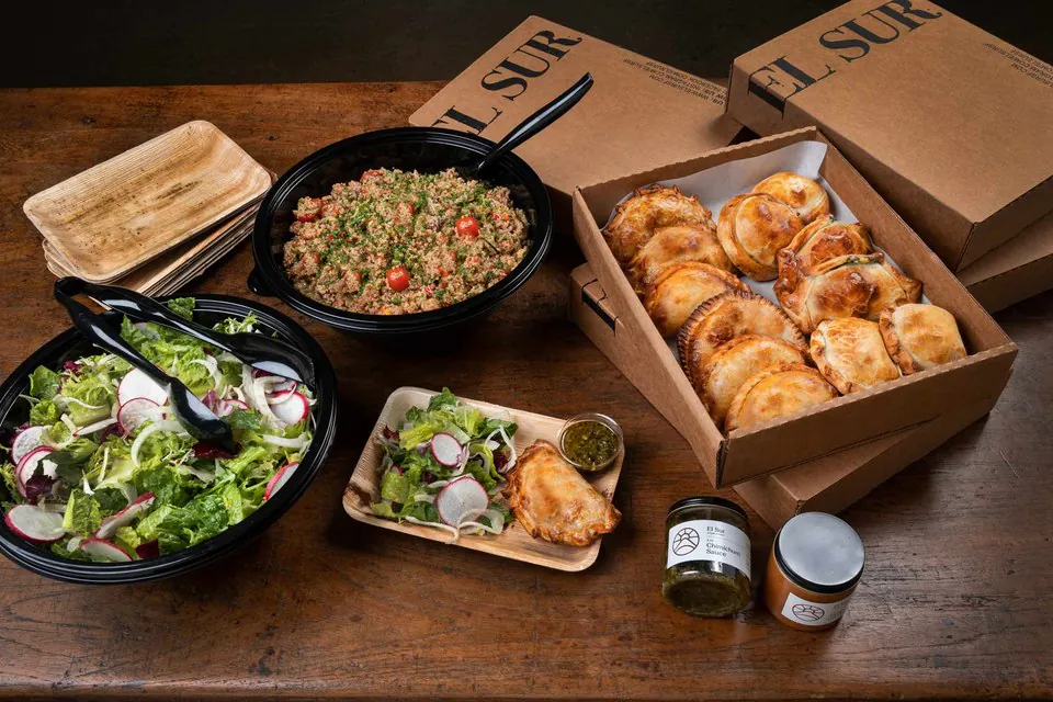 Kraft catering boxes of baked empanadas beside two salads, palm-leaf plates and a jar of chimichurri on a wooden table